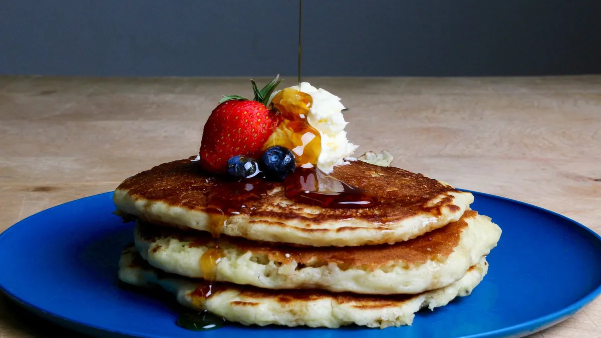 A stack of buttermilk pancakes with maple syrup pouring over and fresh berries in bright breakfast light.